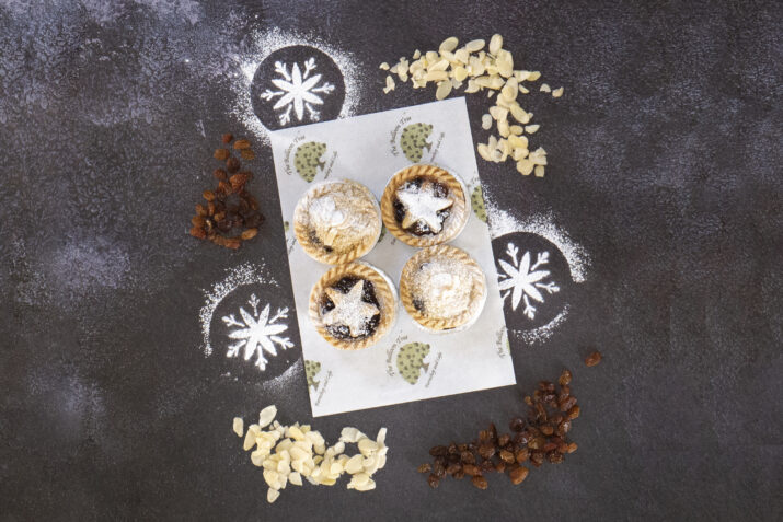 Four homemade mince pies surrounded by flaked almonds, sultanas and icing sugar snowflakes