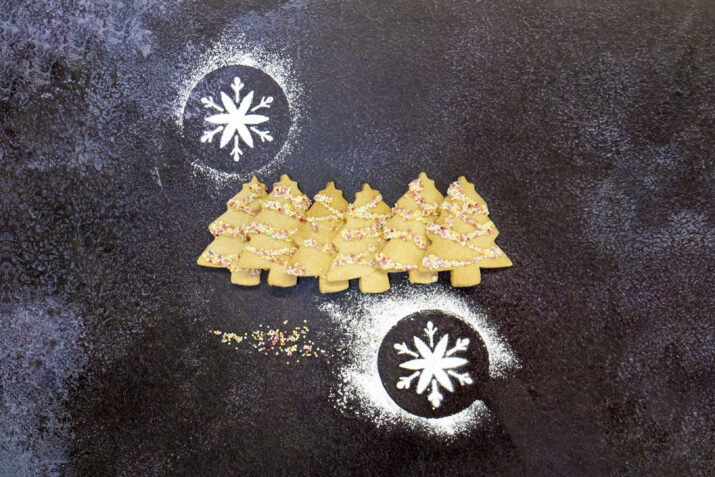 Christmas tree shaped shortbreads surrounded by icing sugar snowflakes